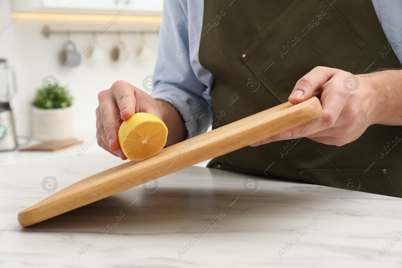 Photo of Man rubbing wooden cutting board with lemon at white table in kitchen, closeup