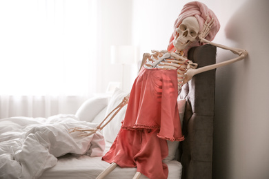 Photo of Human skeleton in silk pajamas and towel sitting on bed indoors