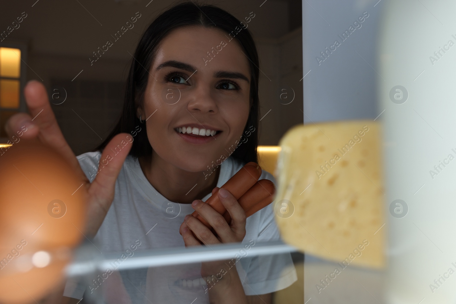 Photo of Young woman taking sausages out of refrigerator in kitchen at night, view from inside