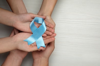 People holding light blue awareness ribbon at white wooden table, top view. Space for text