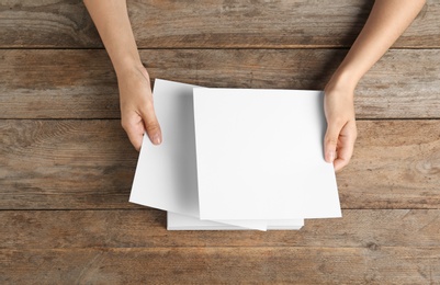 Woman holding blank paper sheets for brochure at wooden table, top view. Mock up