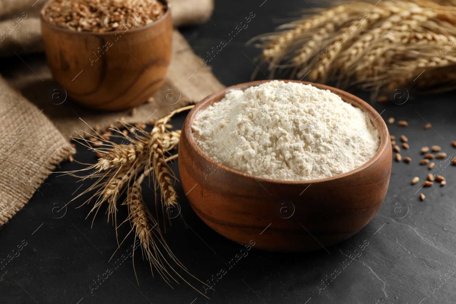 Photo of Wooden bowl of flour and wheat ears on black table