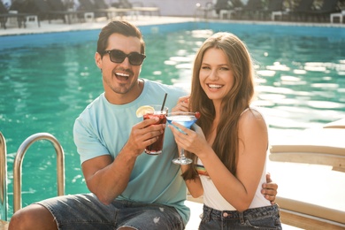 Happy young couple with fresh summer cocktails relaxing near swimming pool