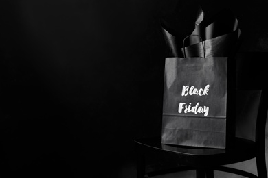 Photo of Shopping bag with words Black Friday on chair against dark background. Space for text
