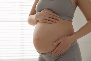 Pregnant young woman touching belly near window indoors, closeup. Space for text