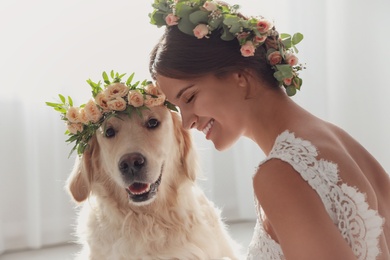 Bride and adorable Golden Retriever wearing wreath made of beautiful flowers indoors