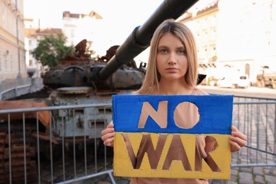 Photo of Sad woman holding poster in colors of Ukrainian flag with words No War near broken tank on city street
