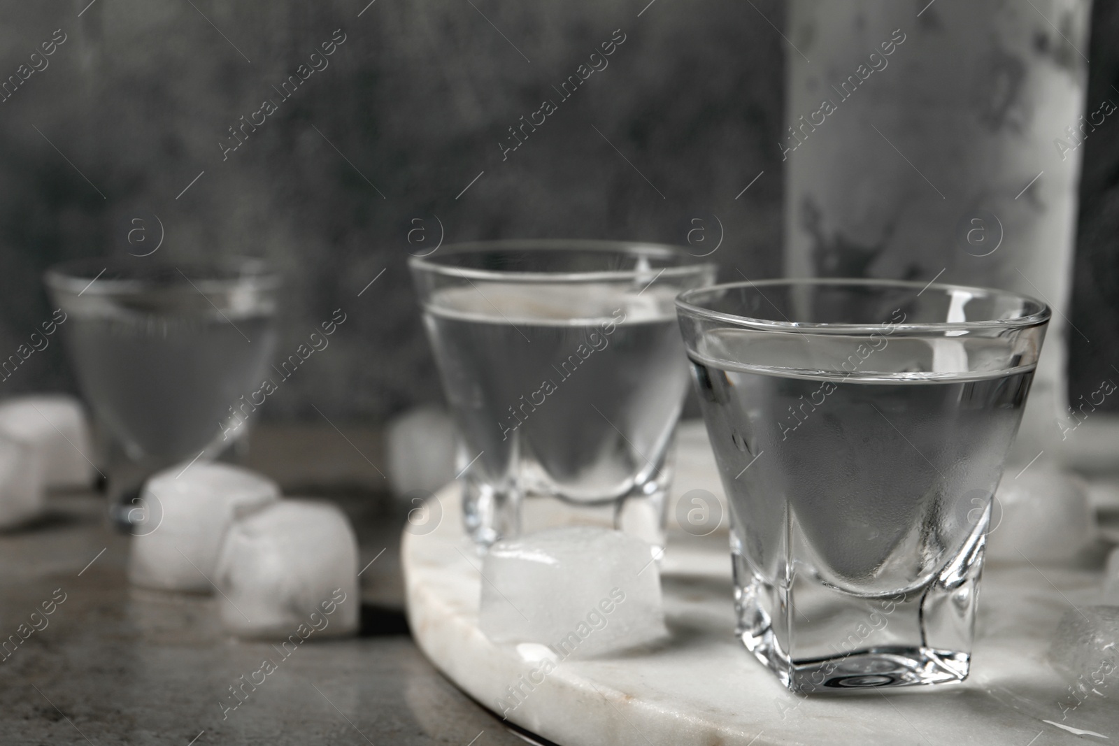 Photo of Shot glasses and bottle of vodka with ice cubes on light grey table, closeup
