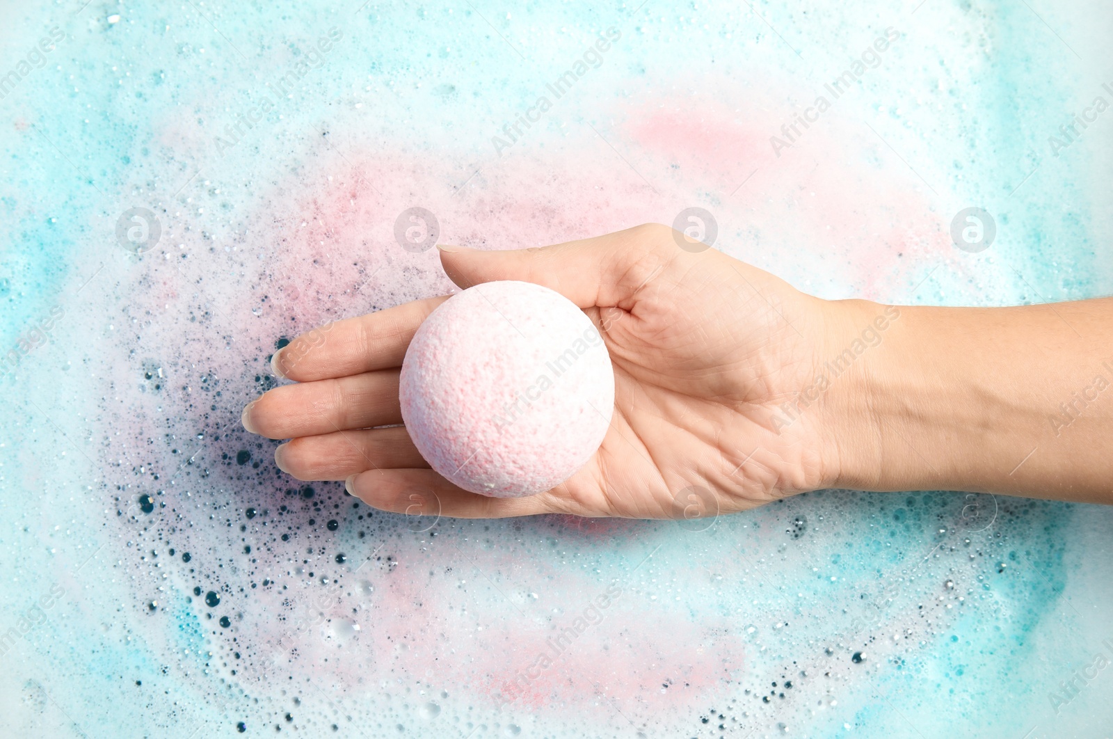 Photo of Woman holding color bath bomb over foam, top view