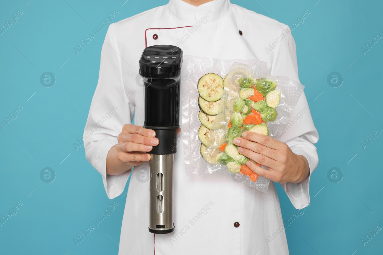 Photo of Chef holding sous vide cooker and vegetables in vacuum packs on light blue background, closeup