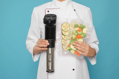 Chef holding sous vide cooker and vegetables in vacuum packs on light blue background, closeup
