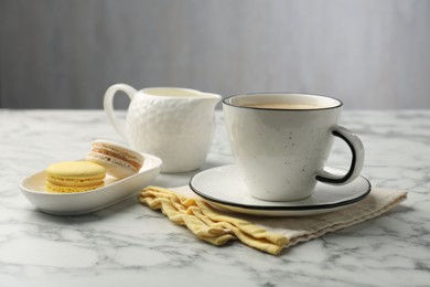 Tasty cappuccino in cup, macarons, pitcher and saucer on white marble table