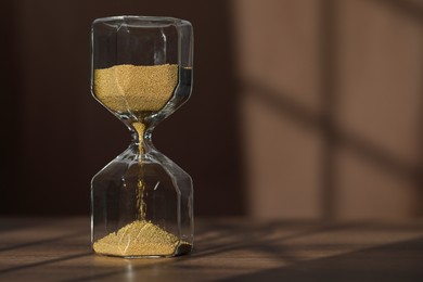 Photo of Hourglass with flowing gold sand on wooden table against brown background, space for text