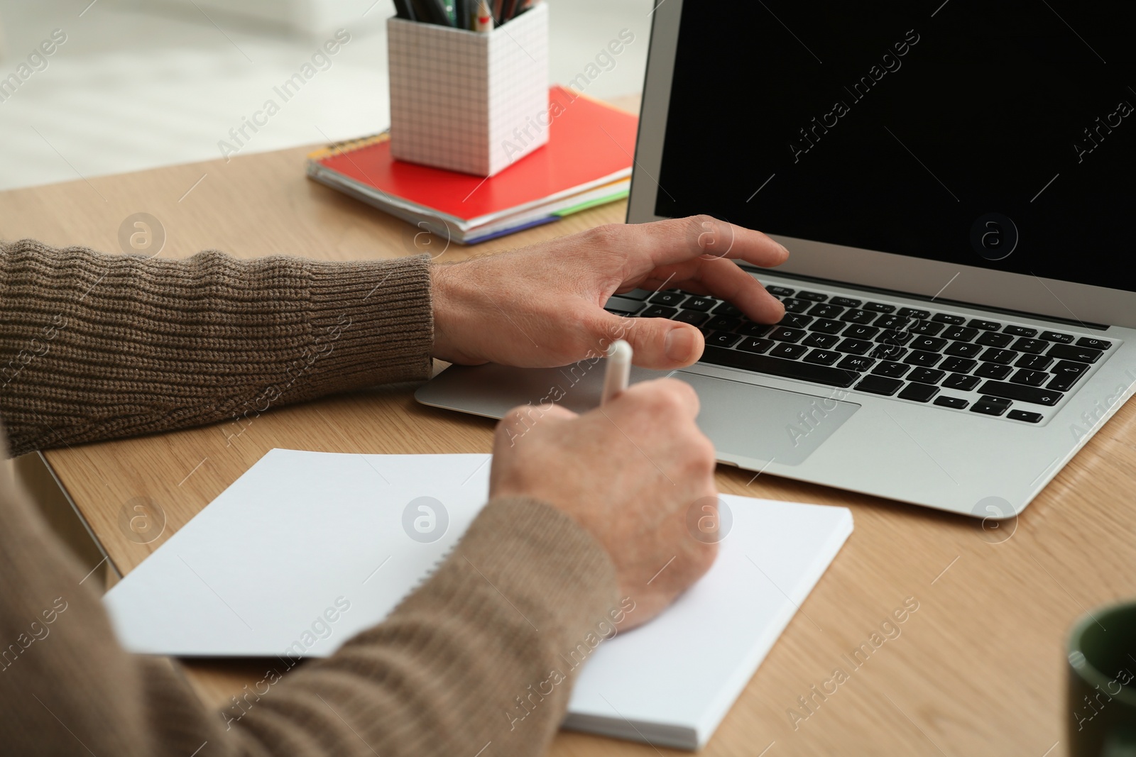 Photo of Man with laptop and notebook learning at wooden table indoors, closeup