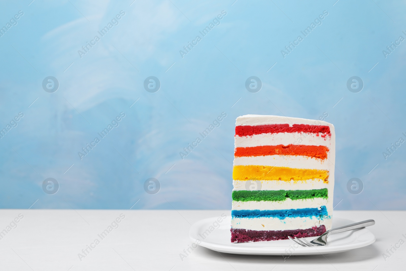 Photo of Plate with slice of delicious rainbow cake on table against color background