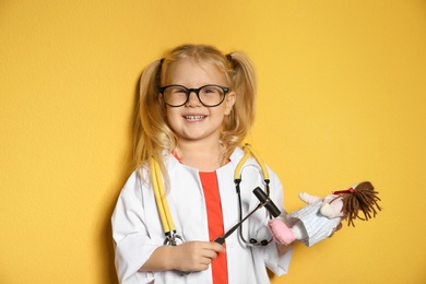 Cute child imagining herself as doctor while playing with reflex hammer and doll on color background
