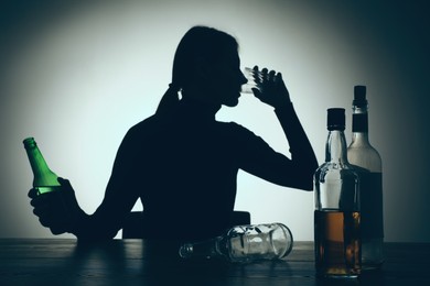 Photo of Alcohol addiction. Silhouette of woman drinking beer on wooden table, backlit