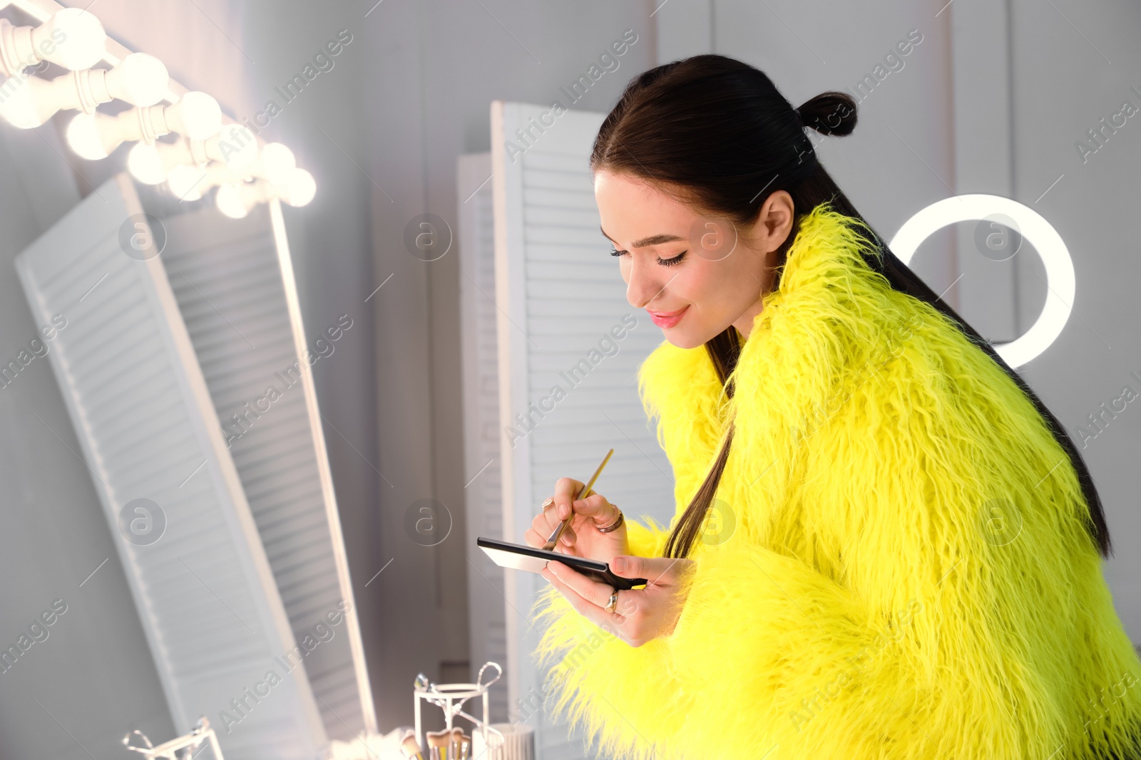 Photo of Young woman applying make up near illuminated mirror indoors