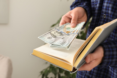 Photo of Man hiding dollar banknotes in book indoors, closeup. Money savings