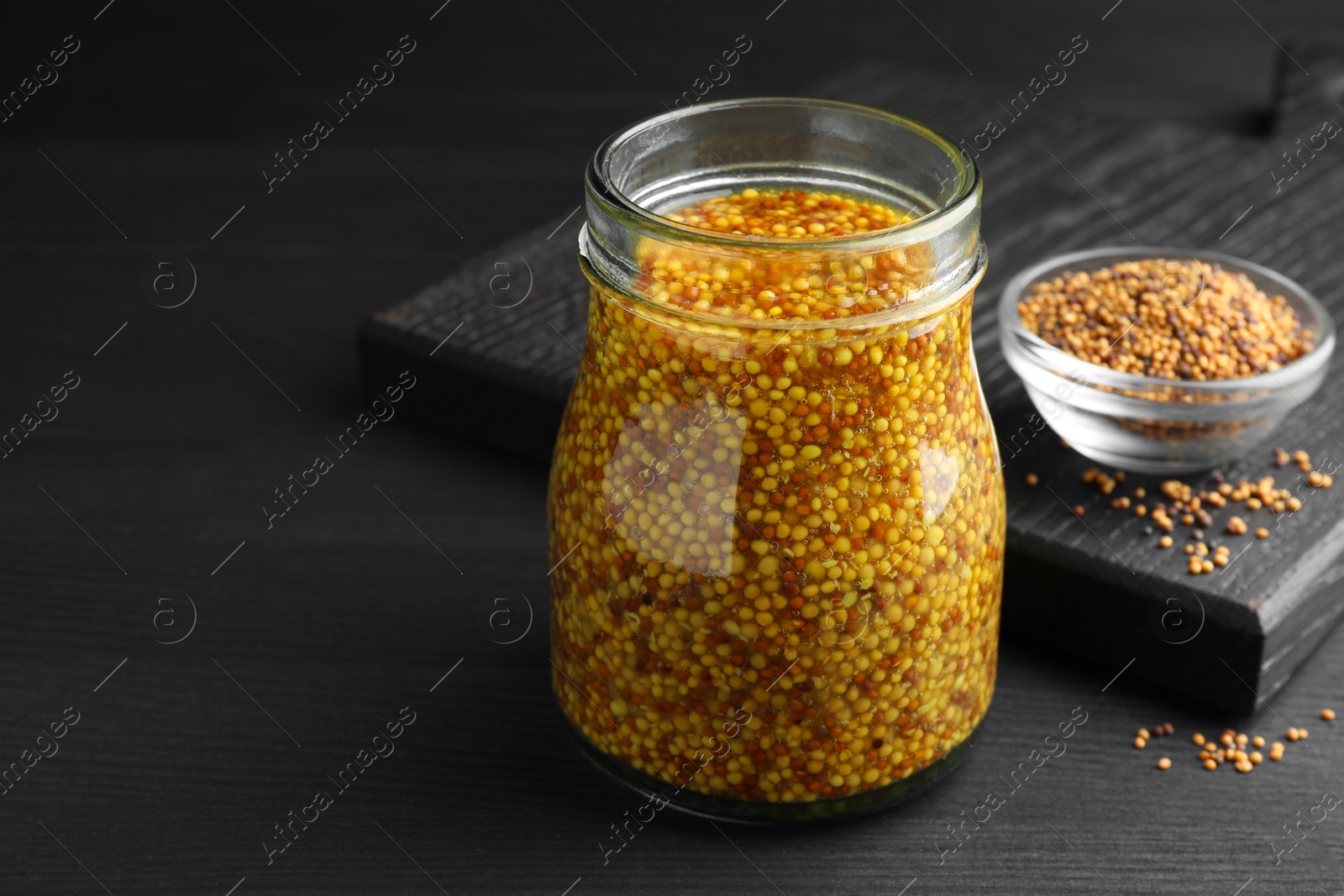Photo of Whole grain mustard in jar and dry seeds on black wooden table. Space for text