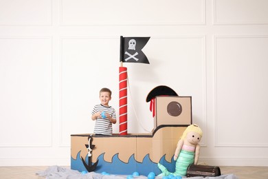 Photo of Cute little boy playing in pirate cardboard ship near white wall indoors