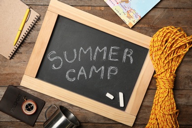 Chalkboard with text SUMMER CAMP and camping equipment on wooden background, flat lay