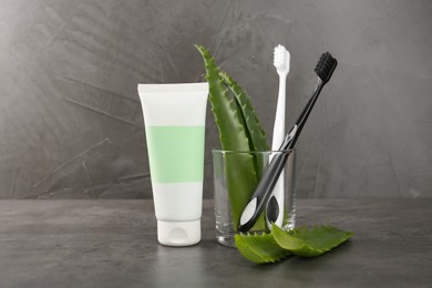 Photo of Aloe vera toothpaste in blank tube, brushes and green leaves on grey table, space for text