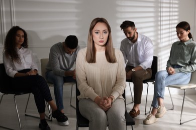 Photo of Unhappy young woman and group of people behind her back indoors. Therapy session