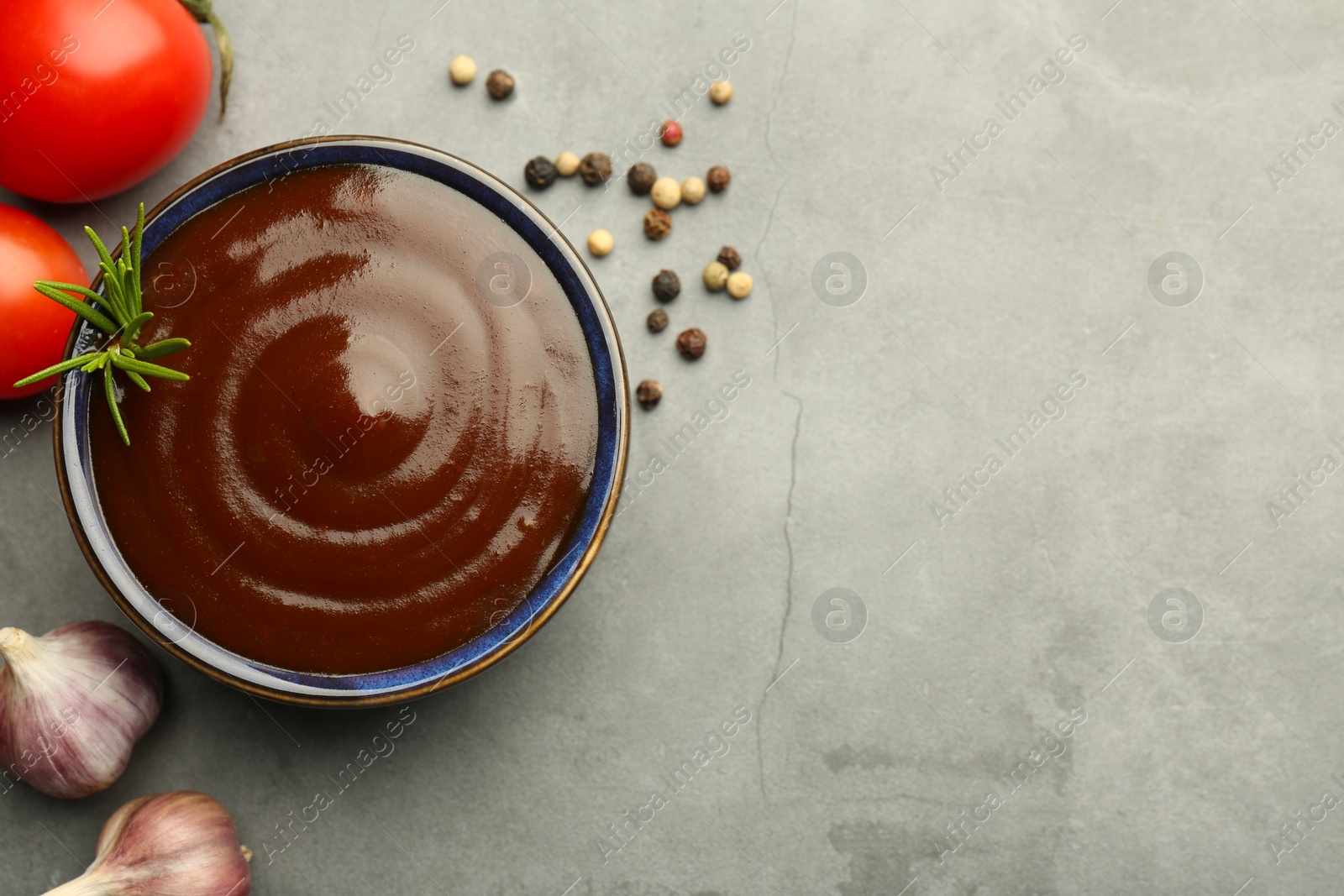 Photo of Tasty barbeque sauce in bowl, rosemary, garlic, tomato and peppercorns on grey textured table, flat lay. Space for text