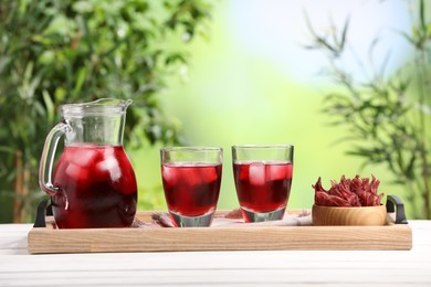 Refreshing hibiscus tea with ice cubes and roselle flowers on white wooden table against blurred green background