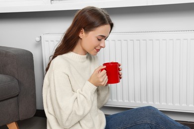 Woman holding cup with hot drink near heating radiator indoors