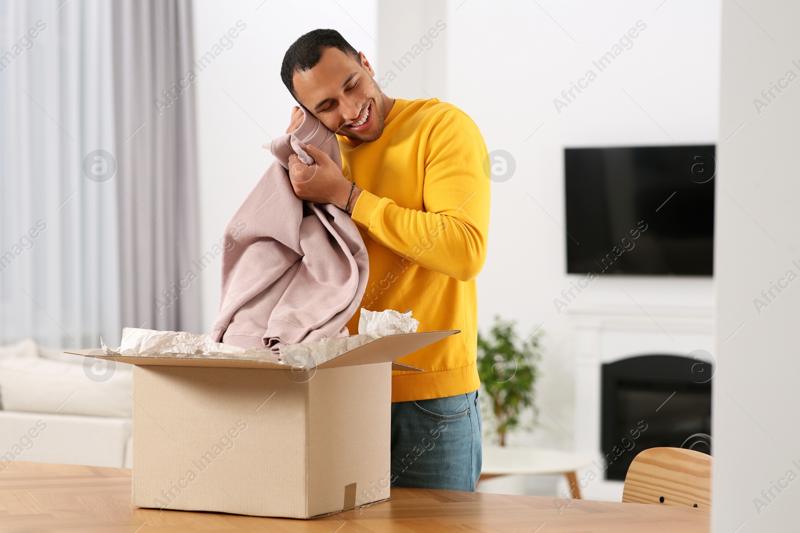 Photo of Happy young man opening parcel at table indoors. Internet shopping