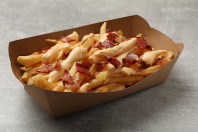 Photo of Tasty potato fries, cheese sauce and bacon in paper container on grey table, closeup