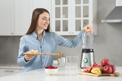 Photo of Beautiful young woman adding banana into blender for tasty smoothie in kitchen