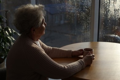 Photo of Elderly woman with drink looking out of window indoors on rainy day, space for text. Loneliness concept