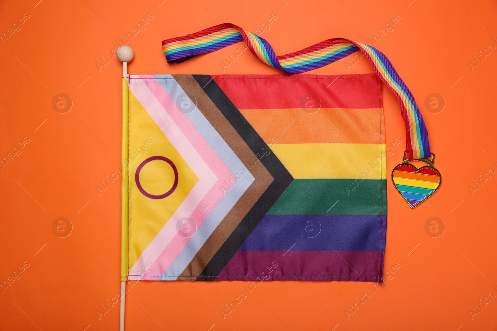Photo of Bright progress flag and rainbow ribbon with heart shaped pendant on orange background, flat lay. LGBT pride