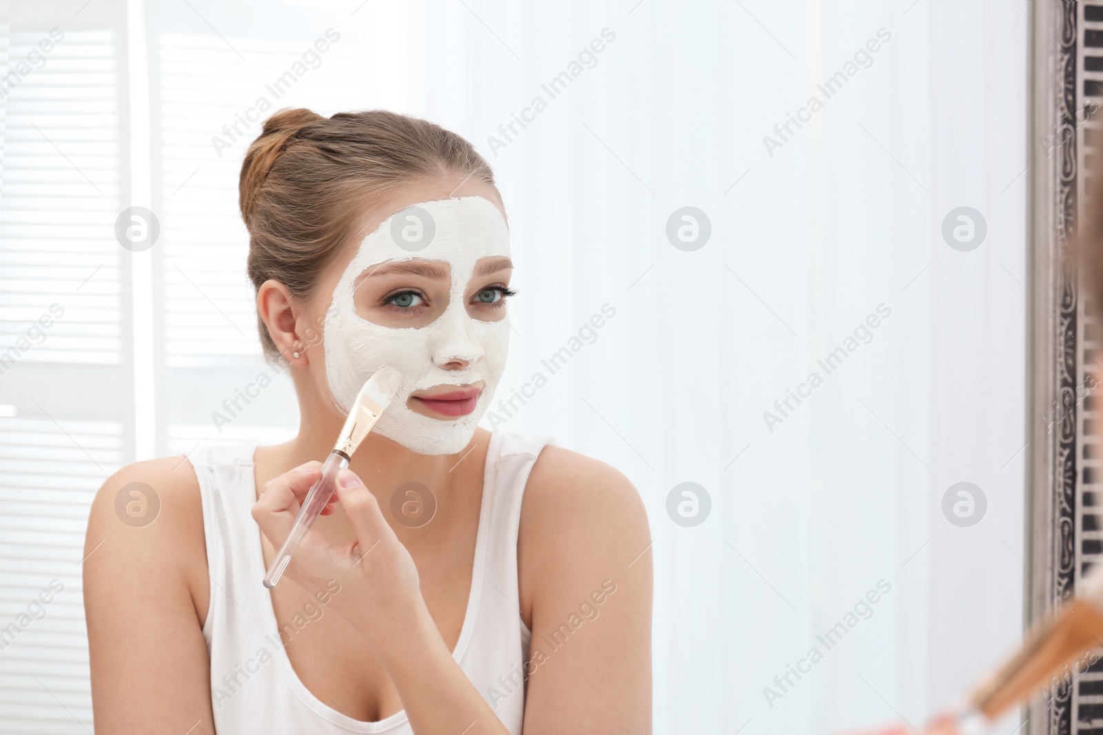 Photo of Beautiful woman applying homemade clay mask on her face at mirror indoors