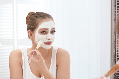 Beautiful woman applying homemade clay mask on her face at mirror indoors