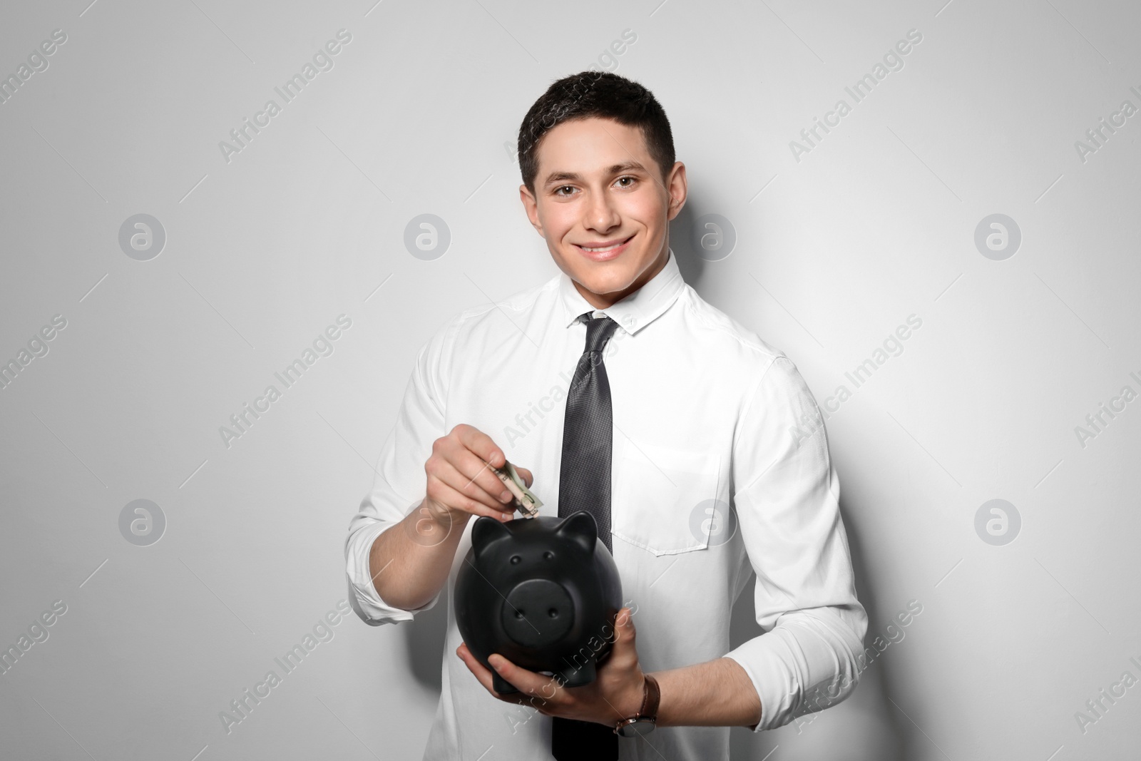 Photo of Young businessman putting money into piggy bank on light background