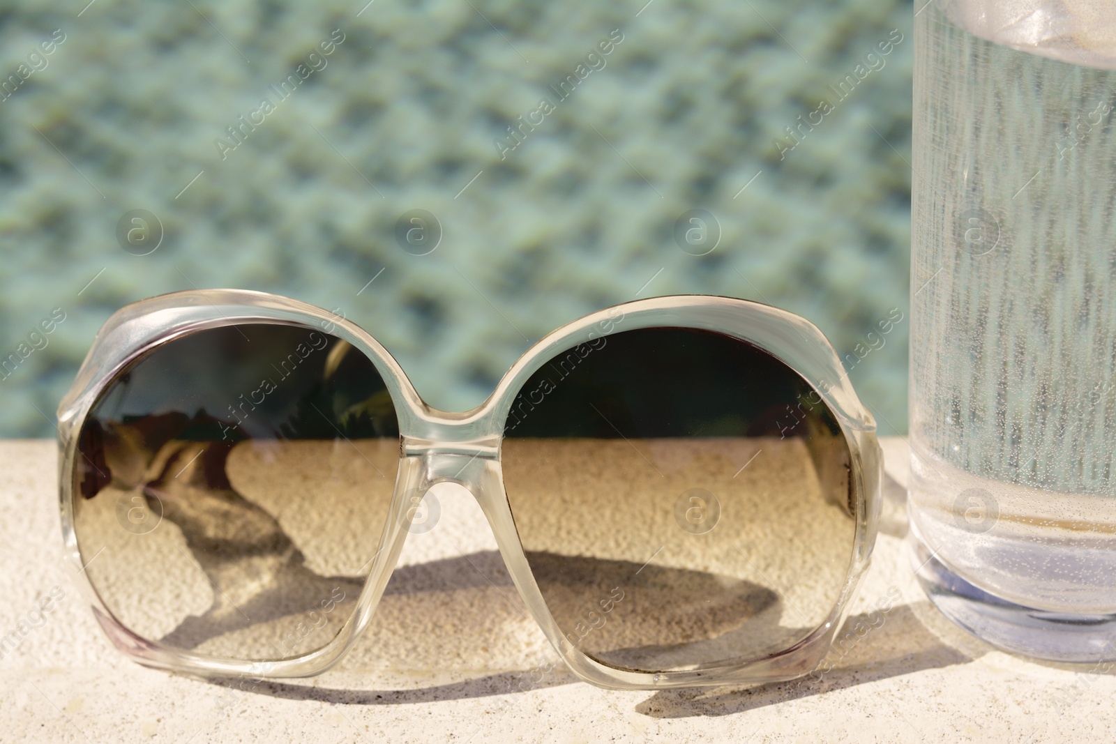 Photo of Stylish sunglasses and glass of water near outdoor swimming pool on sunny day, closeup