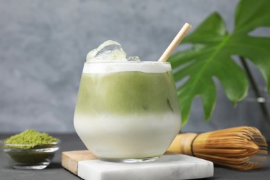 Photo of Glass of tasty iced matcha latte, bamboo whisk and powder on grey wooden table, closeup