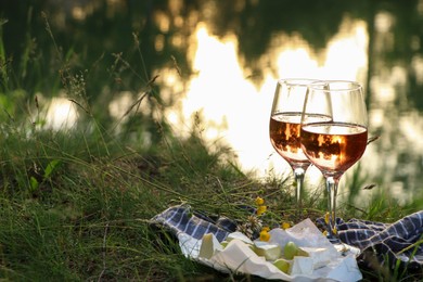 Glasses of delicious rose wine, cheese and grapes on picnic blanket near lake, space for text