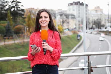 Photo of Young female journalist with microphone and smartphone working on city street. Space for text