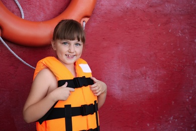 Little girl wearing orange life vest near red wall with safety ring. Space for text