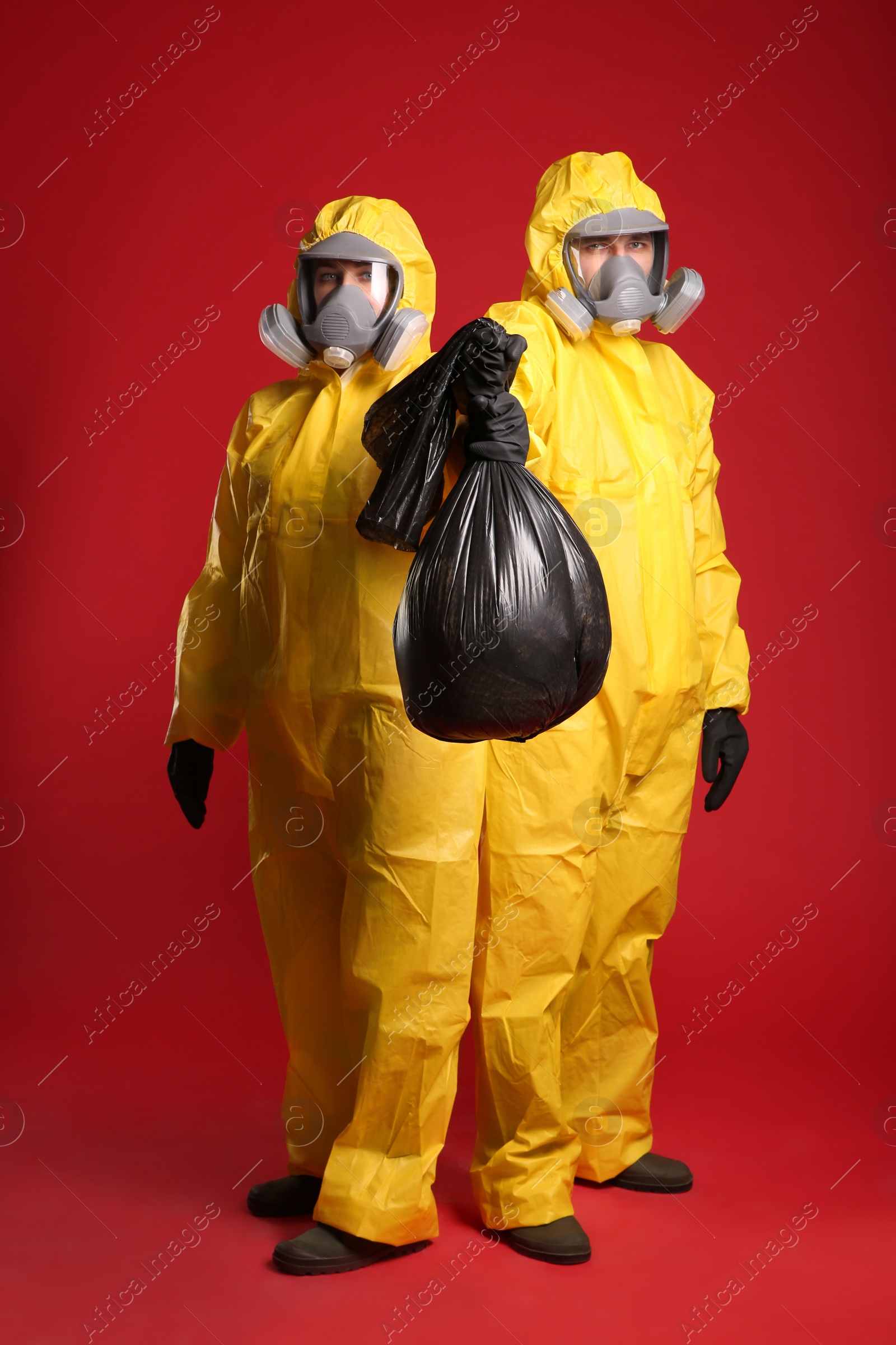 Photo of Man and woman in chemical protective suits holding trash bag on red background. Virus research