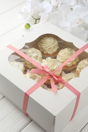 Photo of Tasty cupcakes with vanilla cream in box and orchid flowers on white wooden table, closeup