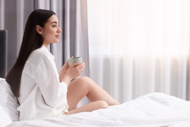 Woman with cup of drink on bed at home, space for text. Lazy morning