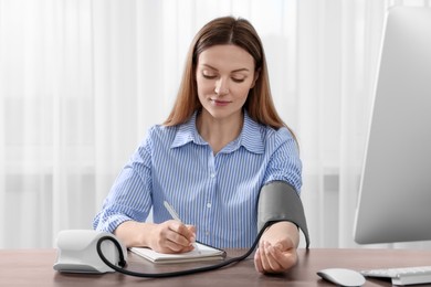 Photo of Woman measuring blood pressure and writing it down into notebook at wooden table in room
