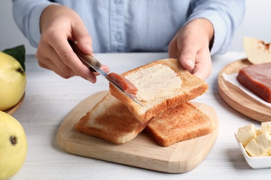 Woman spreading delicious quince paste on toast bread at white wooden table, closeup