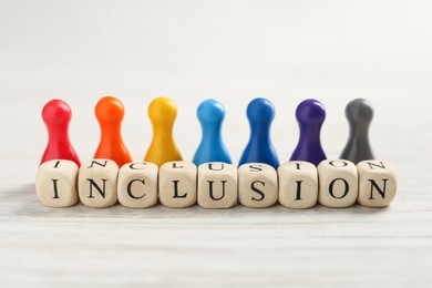 Photo of Colorful pawns and cubes with word Inclusion on white wooden table, closeup